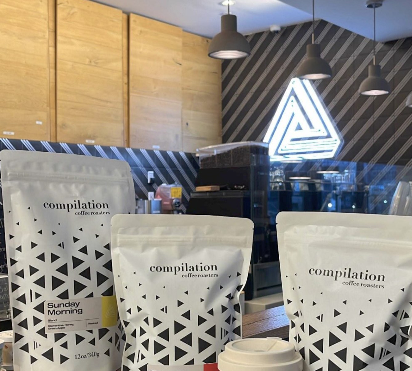Retail bags of Compilation Coffee Roasters beans on the counter beneath the branded triangle logo